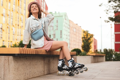 Beautiful young woman with roller skates sitting on bench outdoors, space for text