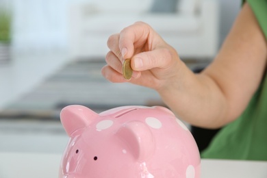 Photo of Mature woman putting money into piggy bank indoors, closeup