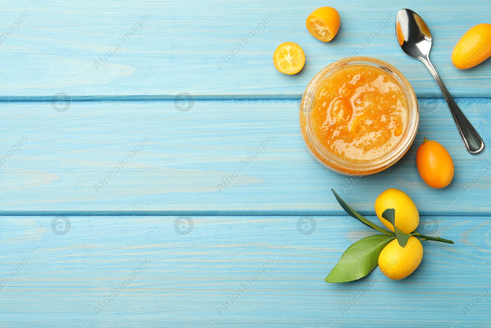 Photo of Jar of delicious kumquat jam, spoon and fresh fruits on light blue wooden table, flat lay. Space for text