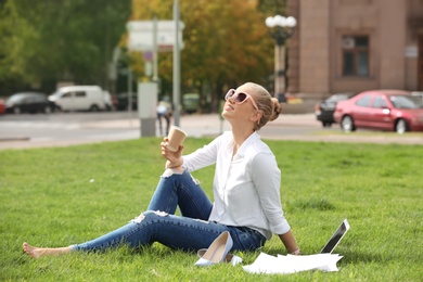 Young woman with cup of coffee sitting on green lawn in park. Joy in moment