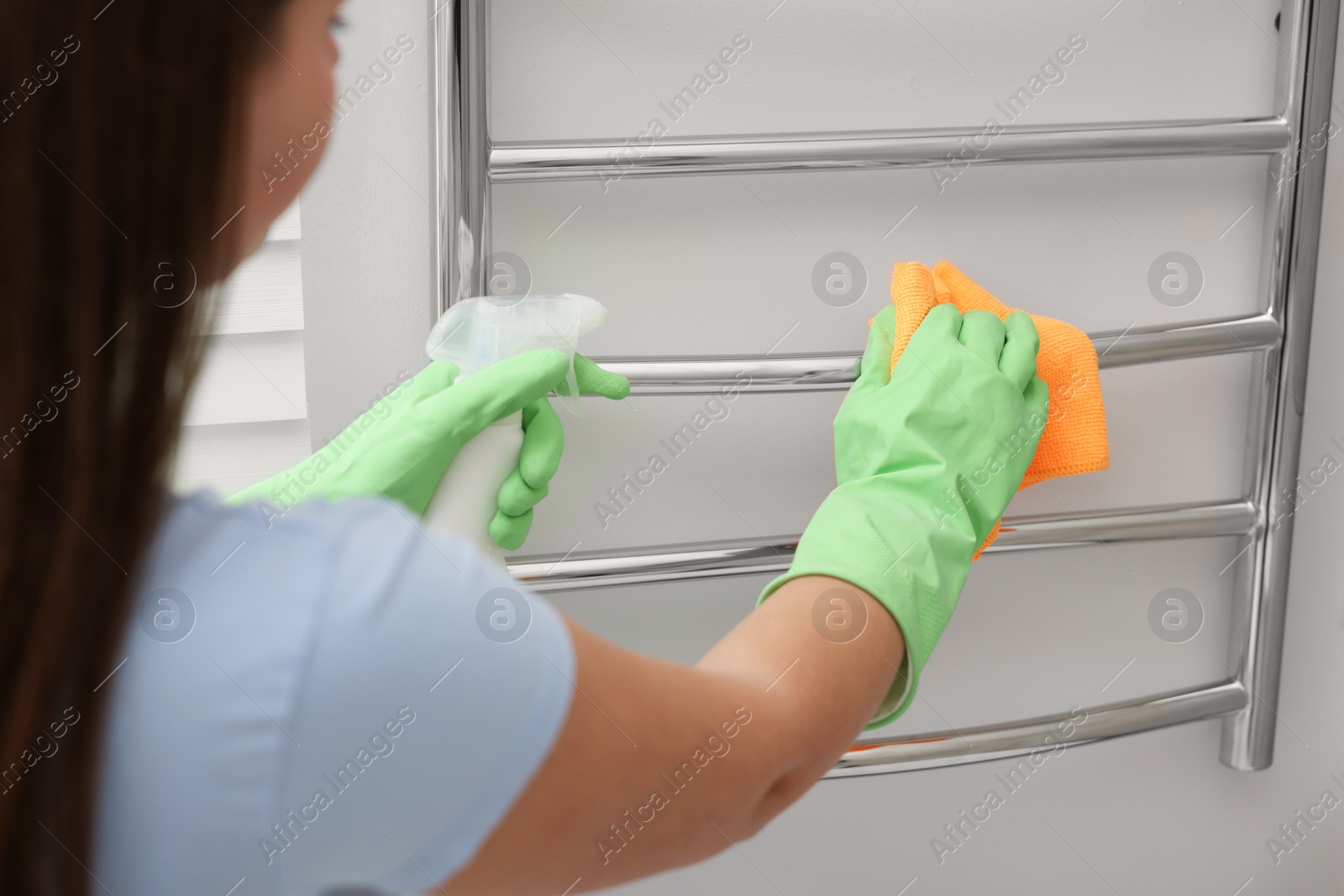 Photo of Woman cleaning heated towel rail with sprayer and rag, closeup