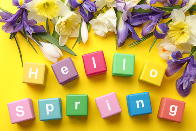 Photo of Colorful cubes with words HELLO SPRING and fresh flowers on yellow background, flat lay