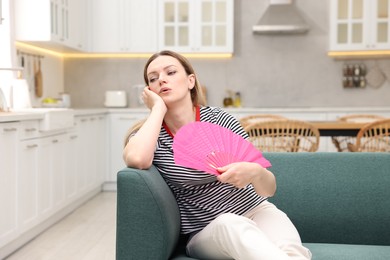 Woman waving pink hand fan to cool herself on sofa at home'