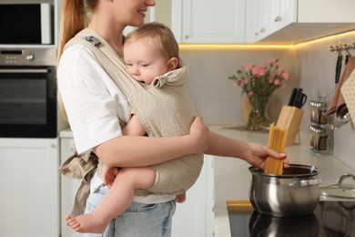 Photo of Mother holding her child in sling (baby carrier) while cooking pasta in kitchen