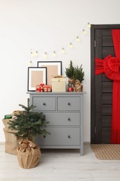 Photo of Wooden door with beautiful red bow near chest of drawers, evergreen trees and gift boxes indoors. Christmas decoration