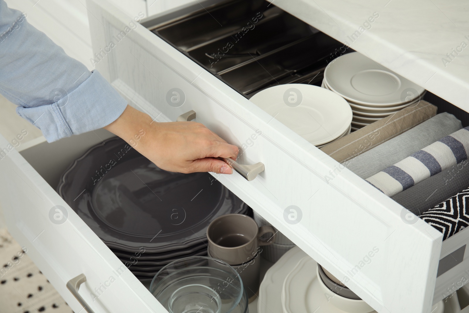 Photo of Woman opening drawer of kitchen cabinet with different dishware and towels, closeup