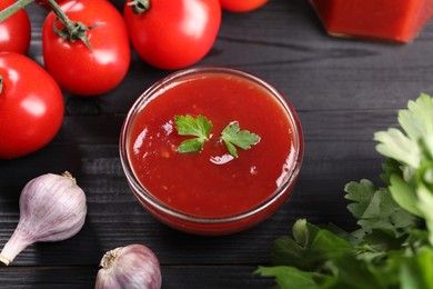 Photo of Delicious ketchup in bowl, garlic, parsley and tomatoes on black wooden table, closeup. Tomato sauce