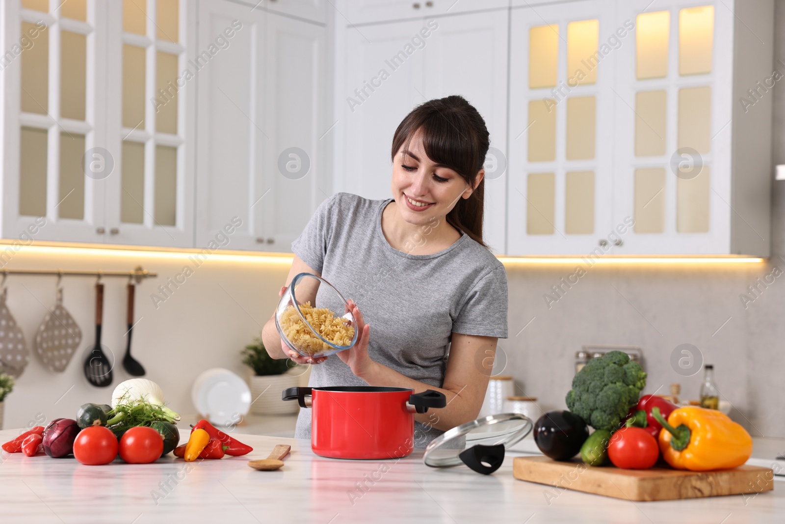 Photo of Happy young housewife adding raw pasta in pot at white marble table in kitchen. Cooking process