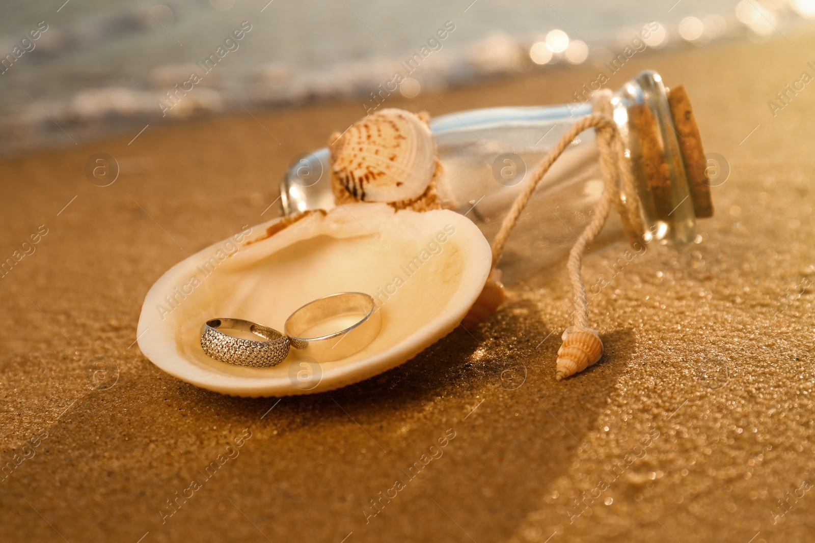 Photo of Shell with gold wedding rings and invitation in glass bottle on sandy beach at sunset, closeup