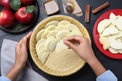 Woman putting apple slices into dish with raw dough at black table, top view. Baking pie