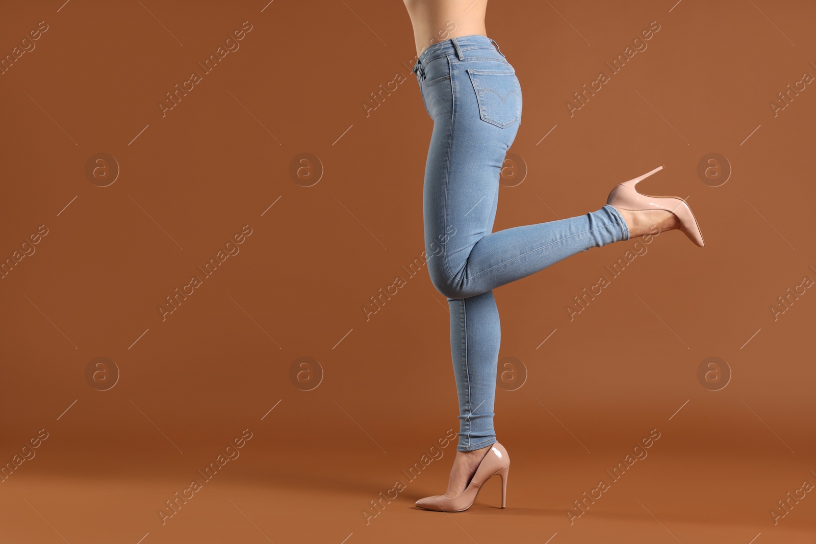 Photo of Woman wearing stylish light blue jeans and high heels shoes on brown background, closeup. Space for text