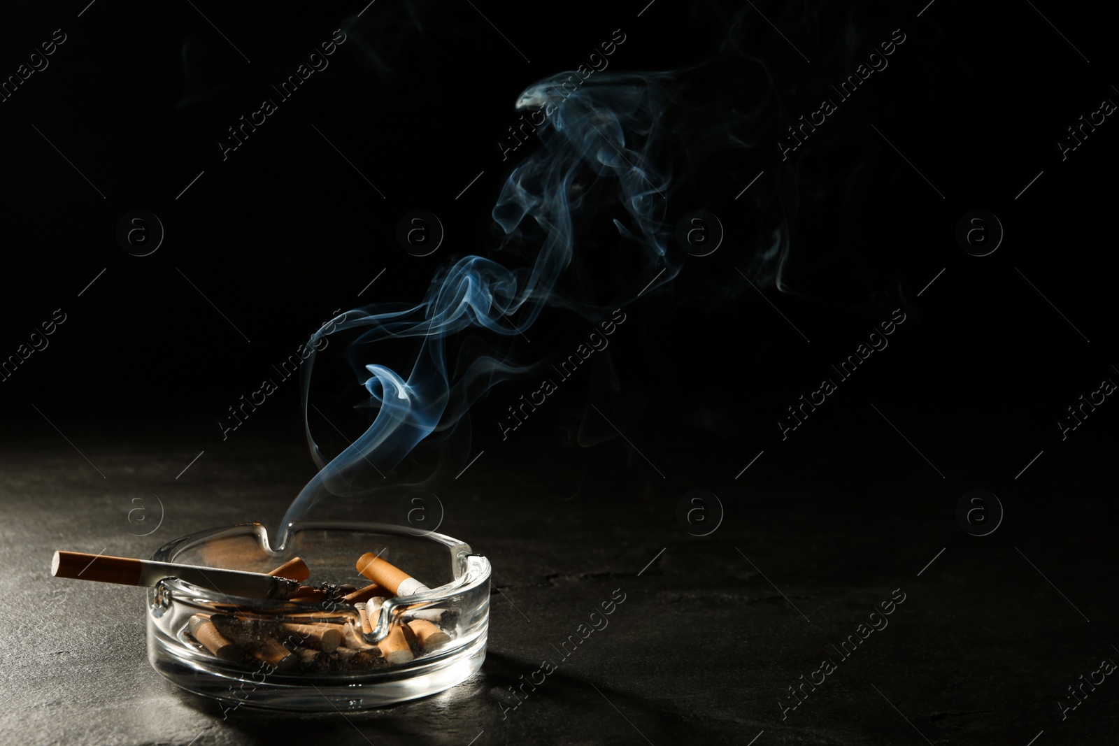 Photo of Smoldering cigarette in glass ashtray on grey table against black background. Space for text