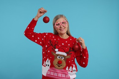 Senior woman in Christmas sweater and funny glasses holding festive baubles on light blue background