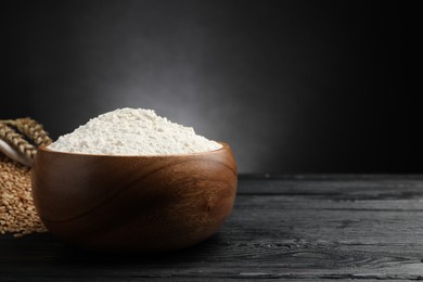 Photo of Wheat flour in bowl, spikes and grains on black wooden table, space for text