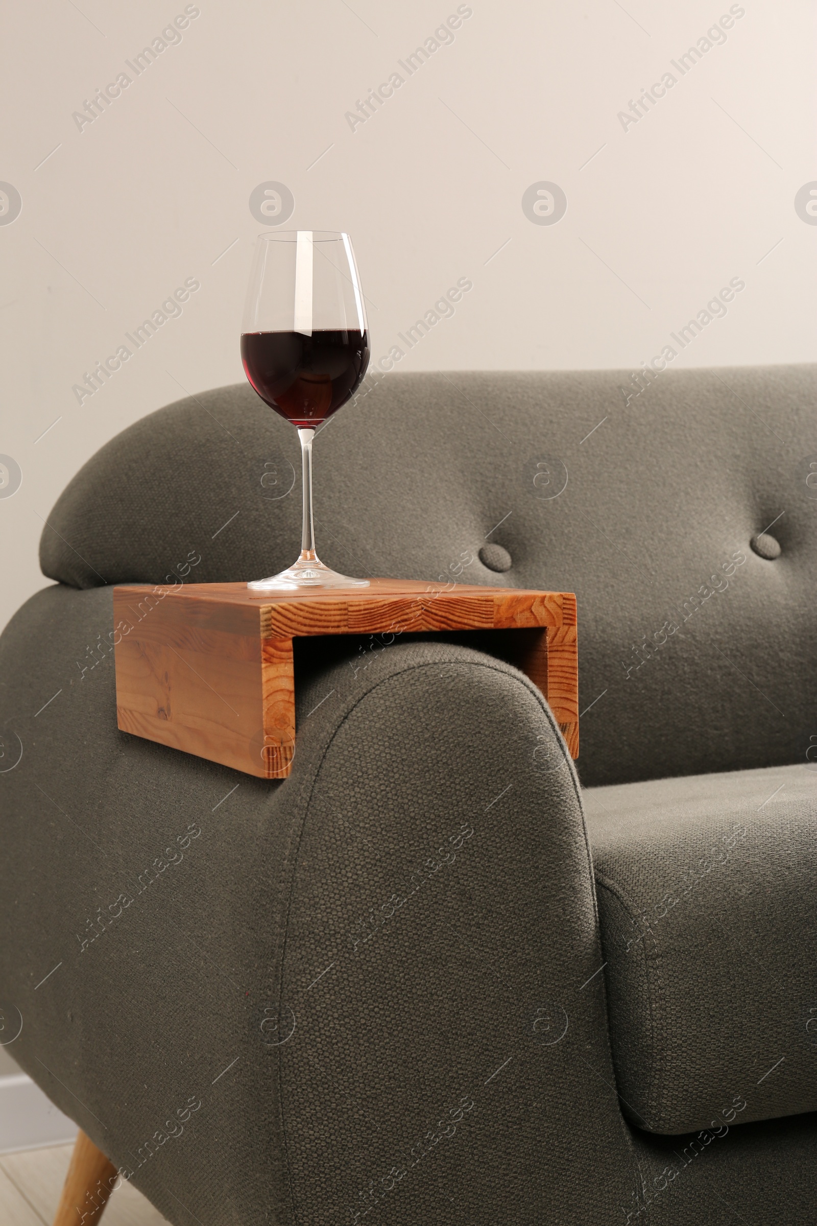 Photo of Glass of red wine on sofa with wooden armrest table in room. Interior element