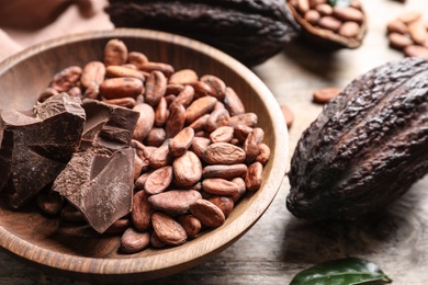Photo of Bowl of cocoa beans and chocolate pieces with pods on wooden table, closeup