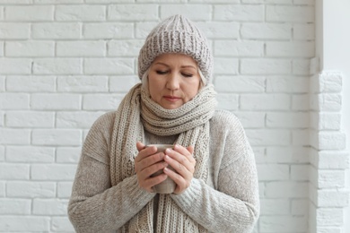 Mature woman in warm clothes suffering from cold on brick background