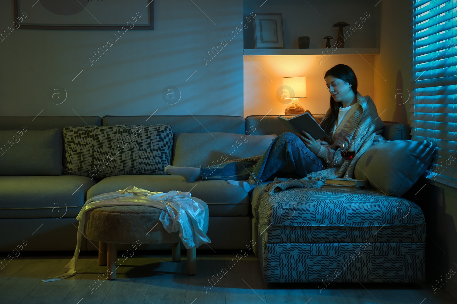 Photo of Woman with glass of wine reading book on couch in room at night