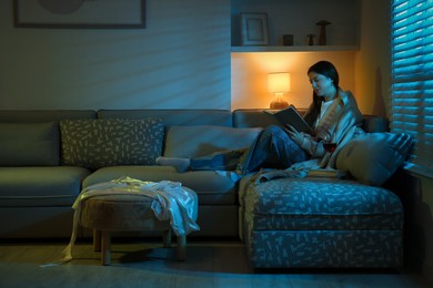 Woman with glass of wine reading book on couch in room at night