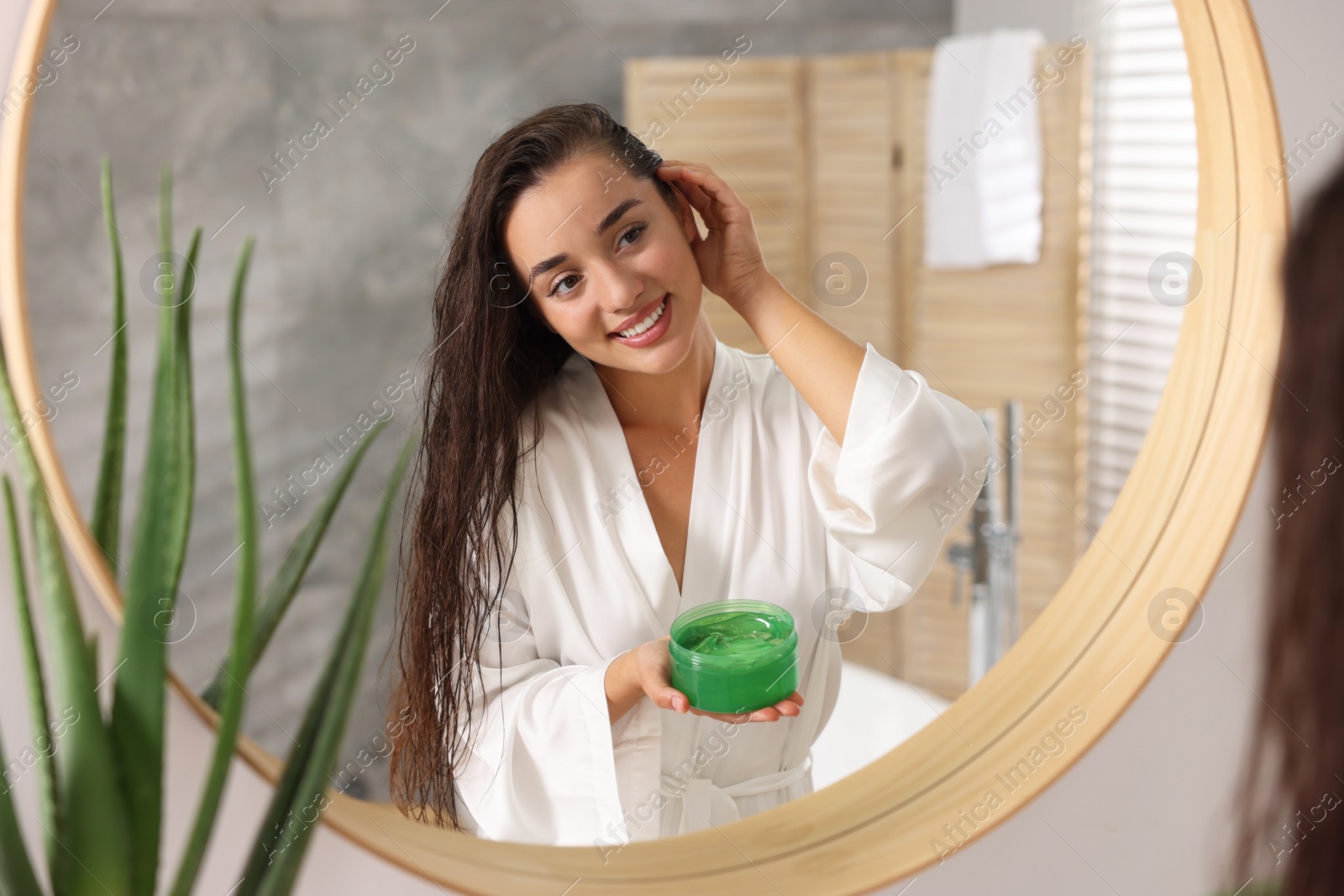 Photo of Young woman applying aloe hair mask near mirror in bathroom