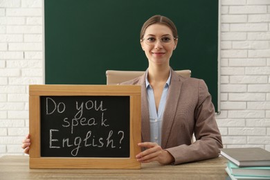 Photo of Teacher holding small chalkboard with inscription Do You Speak English? at table in classroom