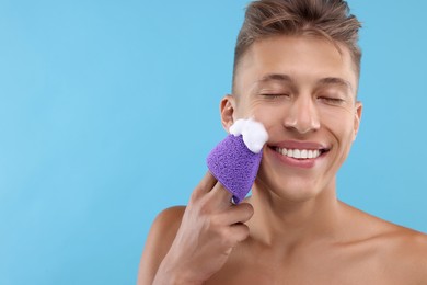 Photo of Happy young man washing his face with sponge on light blue background. Space for text