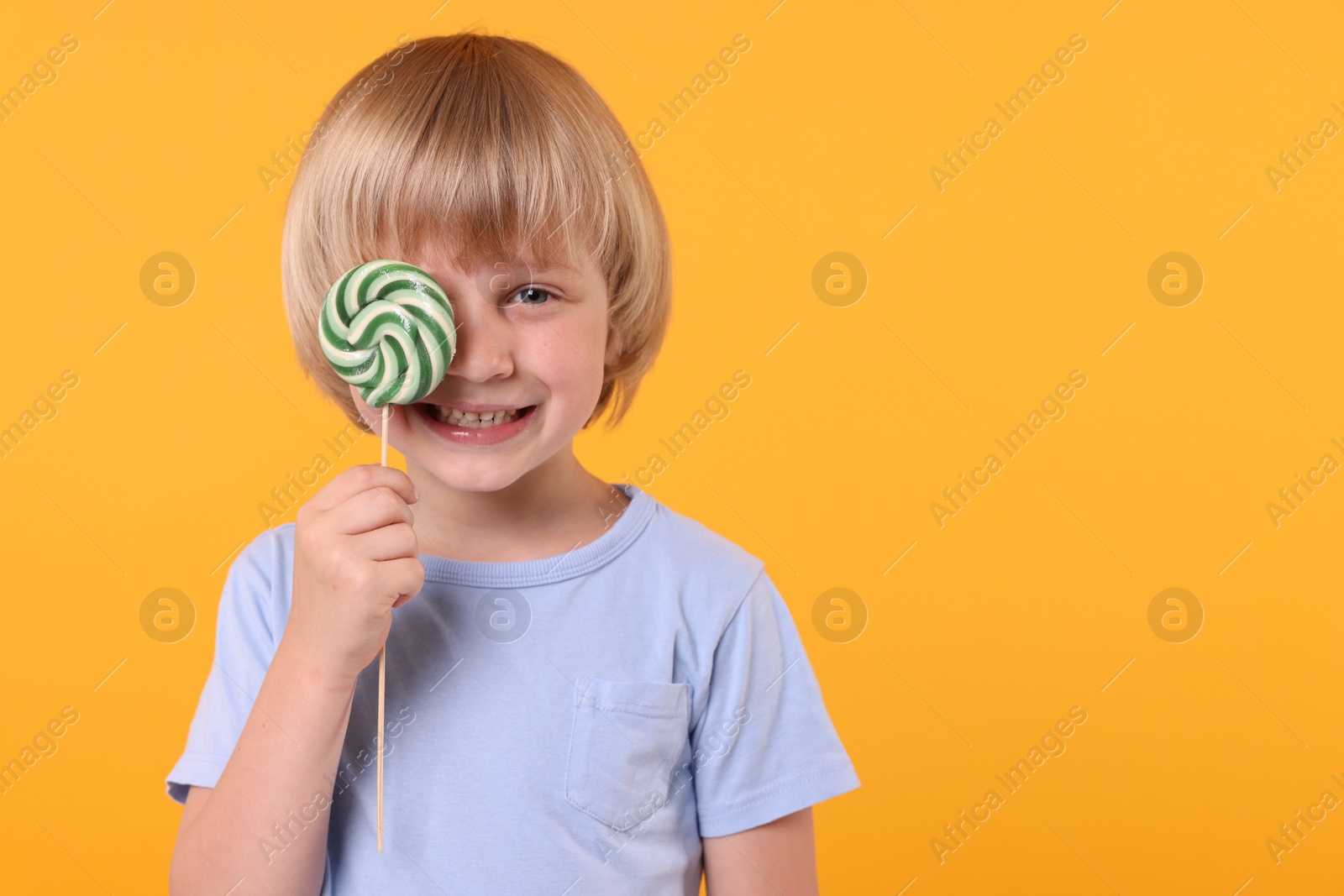 Photo of Happy little boy covering his eye with bright lollipop on orange background, space for text
