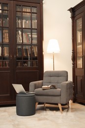 Photo of Comfortable armchair with pouf, laptop and lamp near wooden bookcase in library