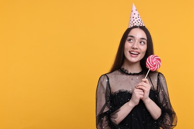 Happy woman in party hat holding lollipop on orange background, space for text