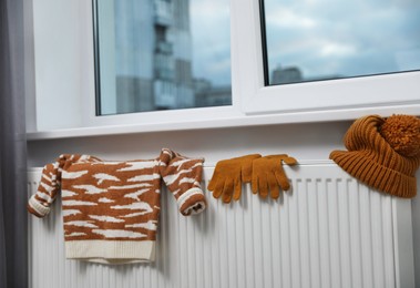 Knitted hat, sweater and gloves on heating radiator near window indoors