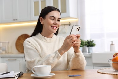 Happy young woman with smartphone shopping online at wooden table in kitchen