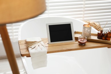 Wooden tray with tablet, cosmetic products and burning candles on bath tub in bathroom