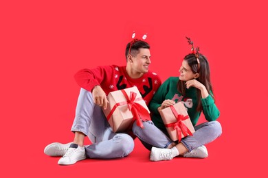 Photo of Beautiful happy couple in Christmas headbands and sweaters sitting with gifts on red background