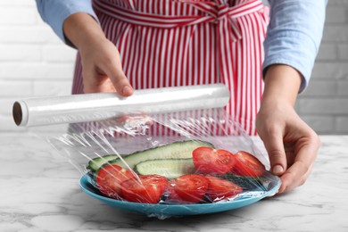 Photo of Woman putting plastic food wrap over plate of fresh vegetables at white marble table indoors, closeup