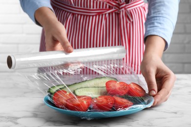 Woman putting plastic food wrap over plate of fresh vegetables at white marble table indoors, closeup