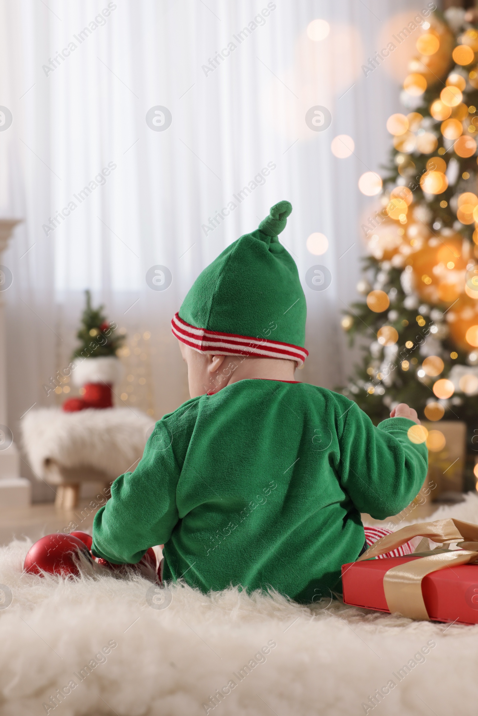 Photo of Baby in cute elf costume near Christmas gift on floor at home, back view