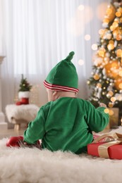 Photo of Baby in cute elf costume near Christmas gift on floor at home, back view
