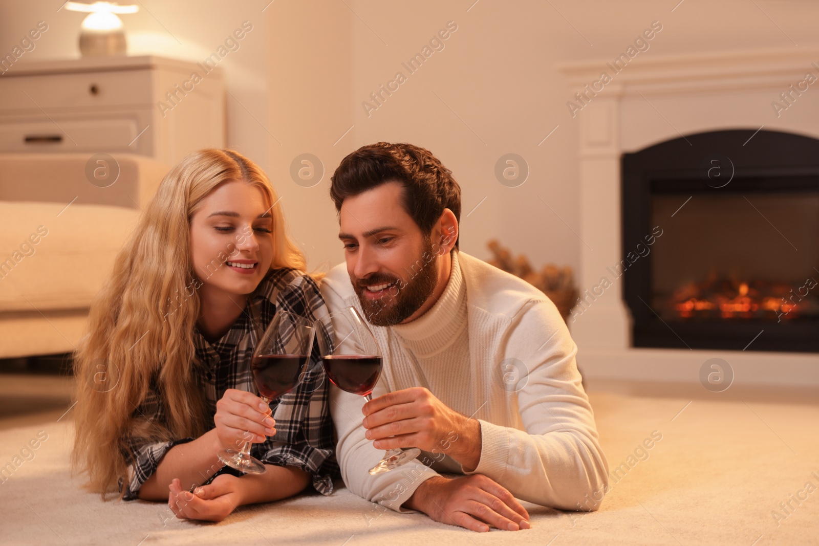 Photo of Lovely couple with glasses of wine spending time together near fireplace at home