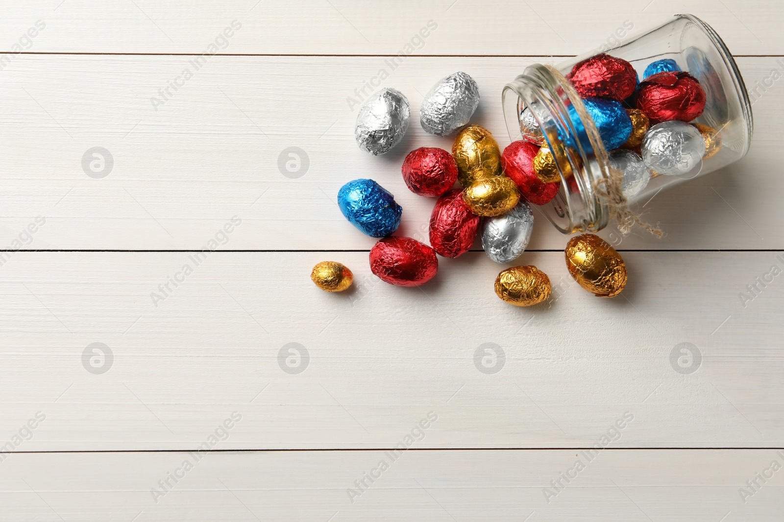 Photo of Overturned glass jar with chocolate eggs wrapped in colorful foil on white wooden table, flat lay. Space for text