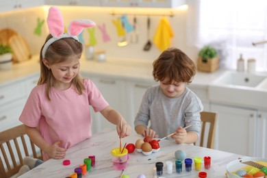 Photo of Easter celebration. Cute children with bunny ears painting eggs at white marble table in kitchen