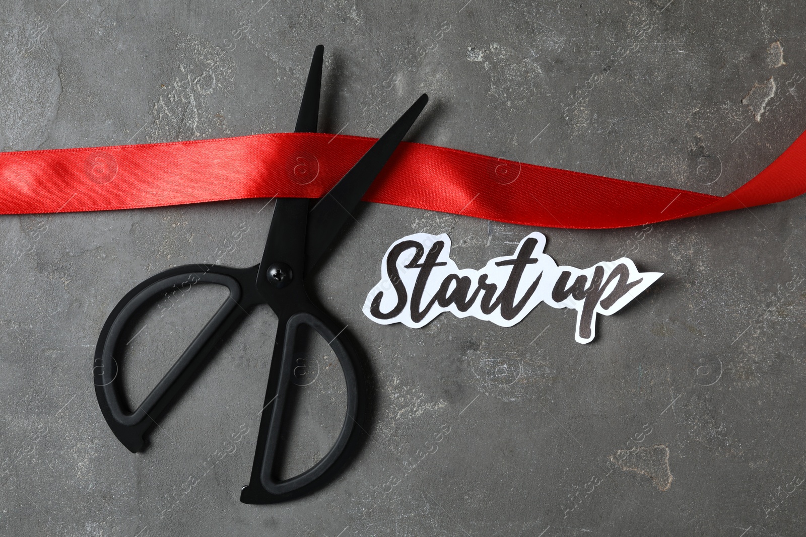 Photo of Sheet of paper with phrase Start Up, red ribbon and scissors on grey table, flat lay