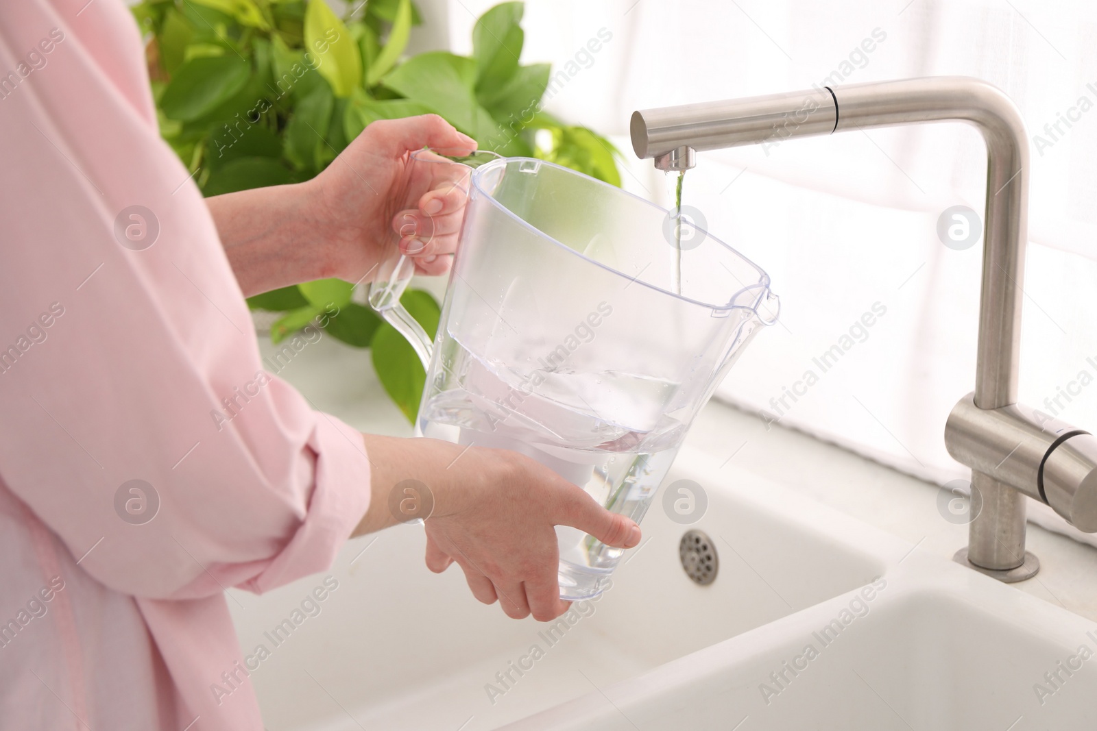 Photo of Woman filling filter jug with water from tap in kitchen, closeup