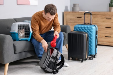 Photo of Travel with pet. Man putting headphones into backpack near carrier with cute cat at home