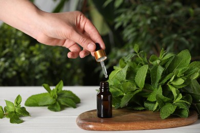 Photo of Woman dripping mint essential oil into bottle at white wooden table, closeup