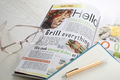 Photo of Open cooking magazine with glasses and notebook on white wooden table, closeup