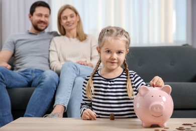 Family budget. Little girl putting coin into piggy bank while her parents watching indoors, selective focus