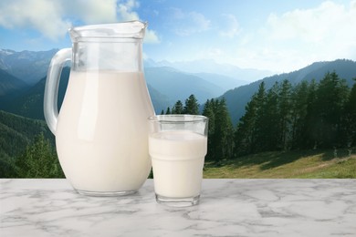 Image of Fresh milk in glass and pitcher on white marble table against mountain landscape, space for text