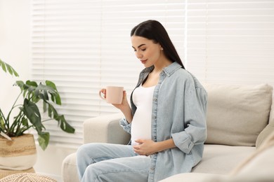 Pregnant woman with cup of drink on sofa at home