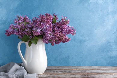 Photo of Jug with beautiful blossoming lilac on table against color background. Spring flowers