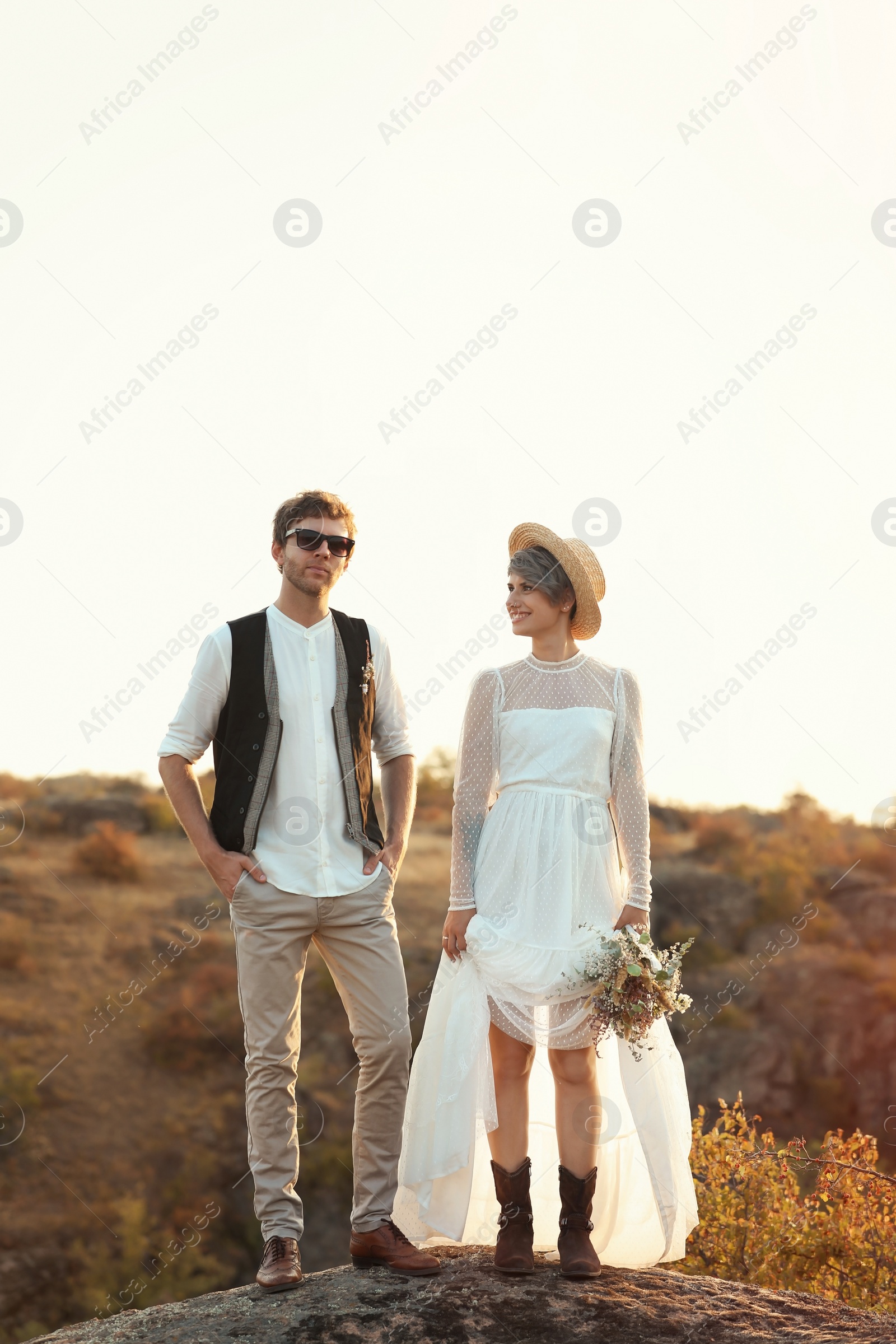 Photo of Happy newlyweds with beautiful field bouquet standing on rock outdoors